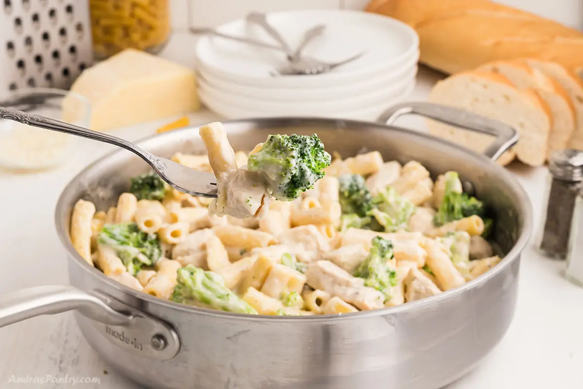 A form taking some creamy pasta out of a deep stainless steel pan with chicken broccoli ziti.
