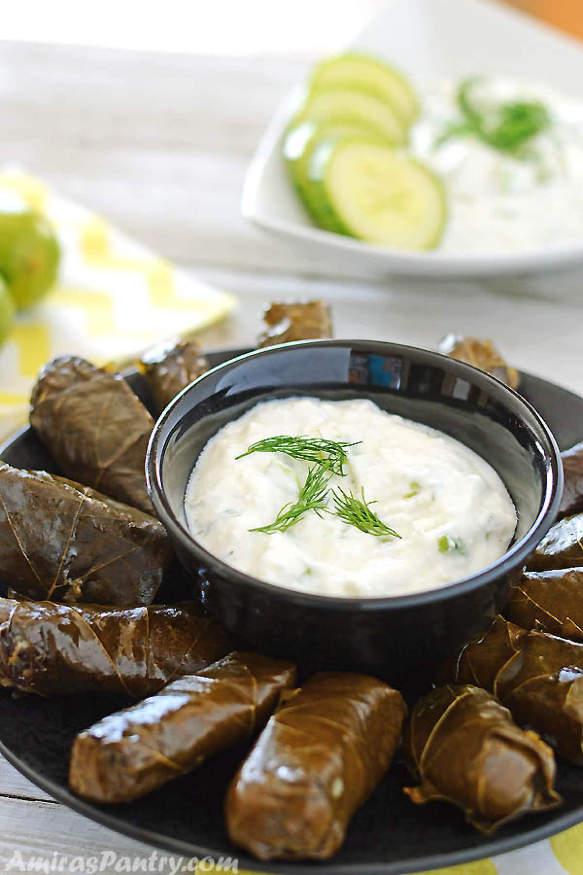 A small black bowl with Greek yogurt sauce sauce surrounded by stuffed grape leaves.