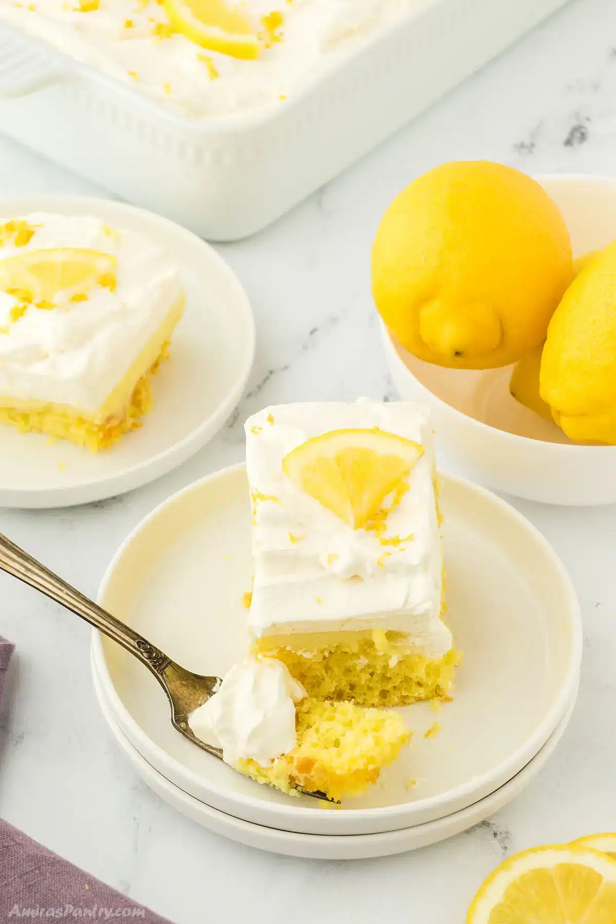 Lemon poke cake pieces on white plates with a bowl of fresh lemon.