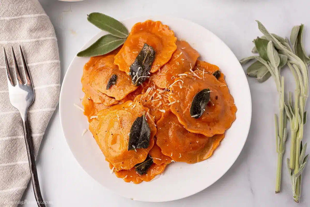 A white plate with a serving of butternut squash ravioli garnished with sage leaves.