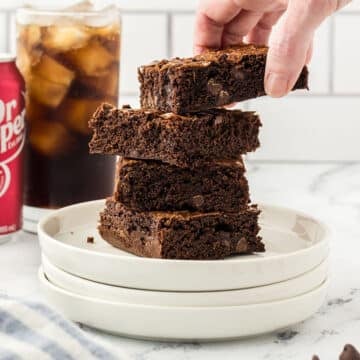 A stack of brownies on a white plate with dr pepper glass in the back.