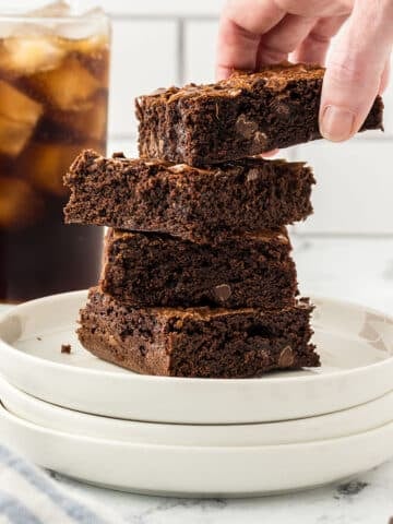 A stack of brownies on a white plate with dr pepper glass in the back.