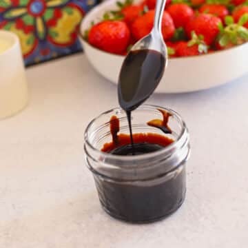 Spoon dripping some balsamic glaze over a glass jar with strawberries in the back.