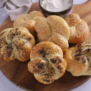 A pile of kaiser rolls on a wooden board with cheese.