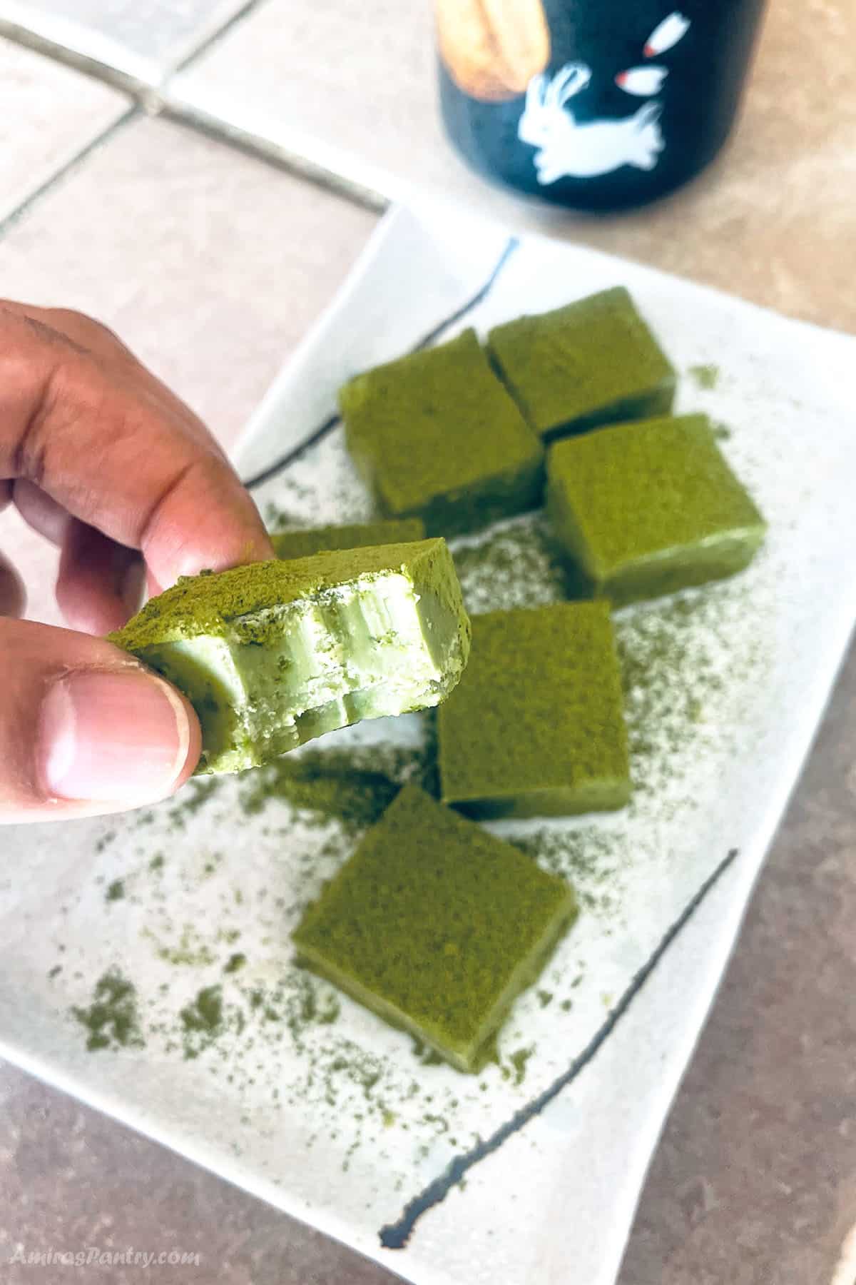 A hand holding a square of matcha chocolate with a bite taken off to show texture.