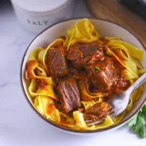 A white bowl with beef cubes on egg noodles.