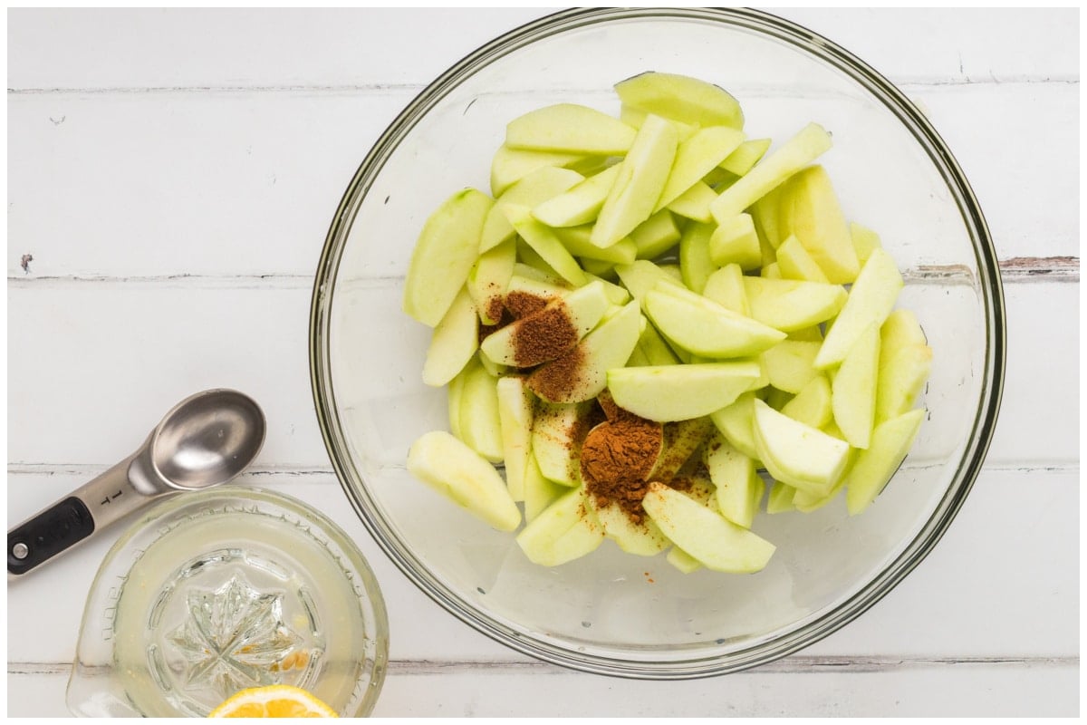 A large glass bowl with silced apples, cinnamon and nutmeg.