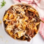An overhead image of a white bowl with beef ragu garnished with parmesan cheese.