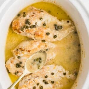 A spoon with piccata sauce over a white slow cooker.