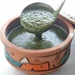 A ladle scooping molokhia from a clay pot on a white table with some pita bread on the side.