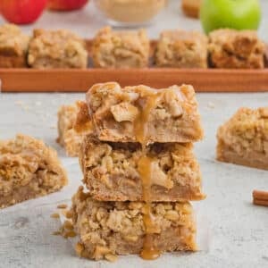 Apple pie bars with caramel sauce dripping and many apples in the back.