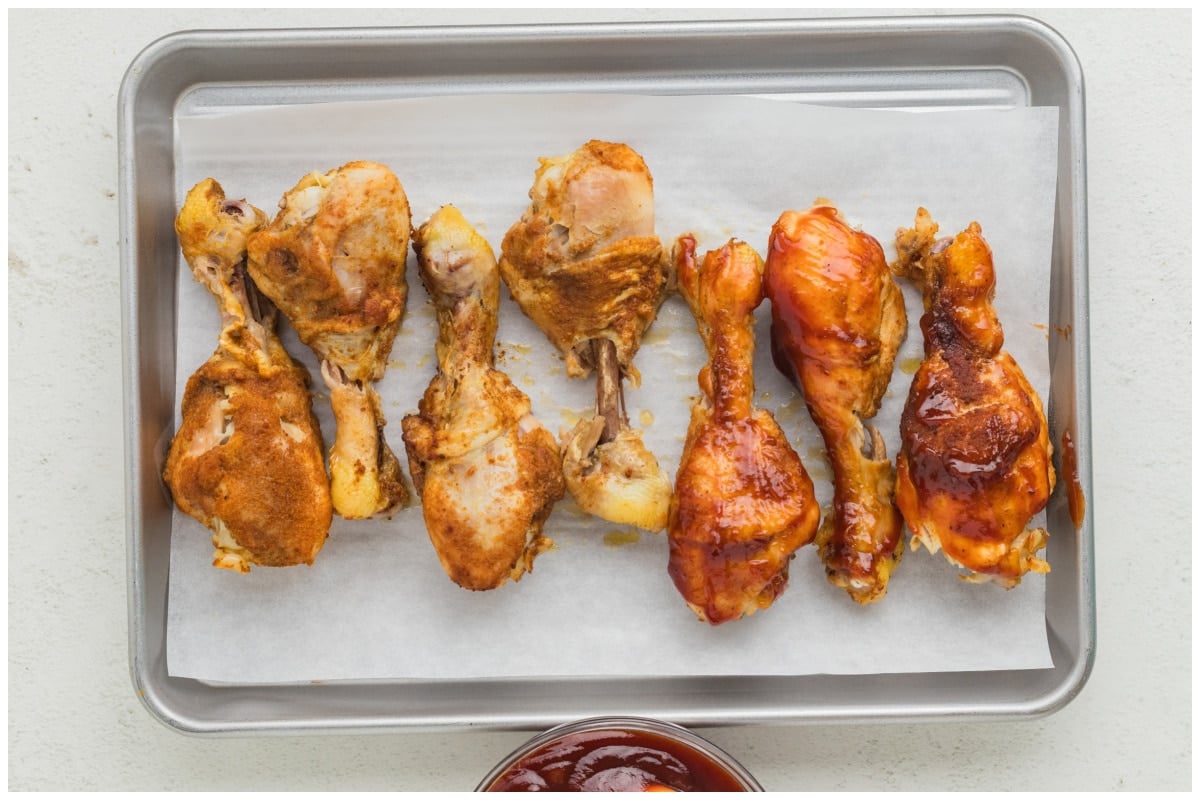 Drumsticks on a baking sheet with half brushed with bbq sauce.