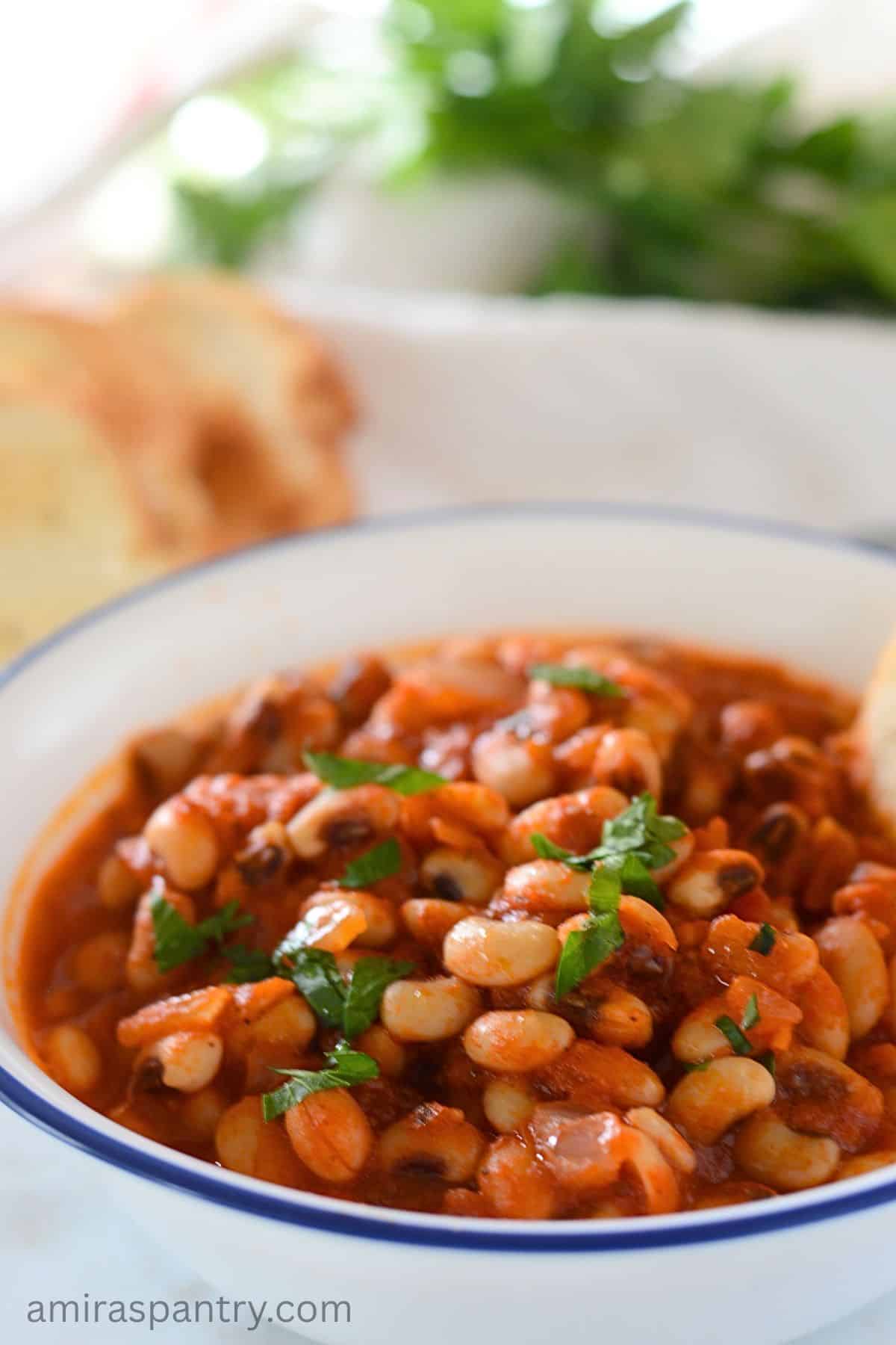 Black eyed peas stew in a white bowl with a dark blue rim. A piece of bread is in the bowl.