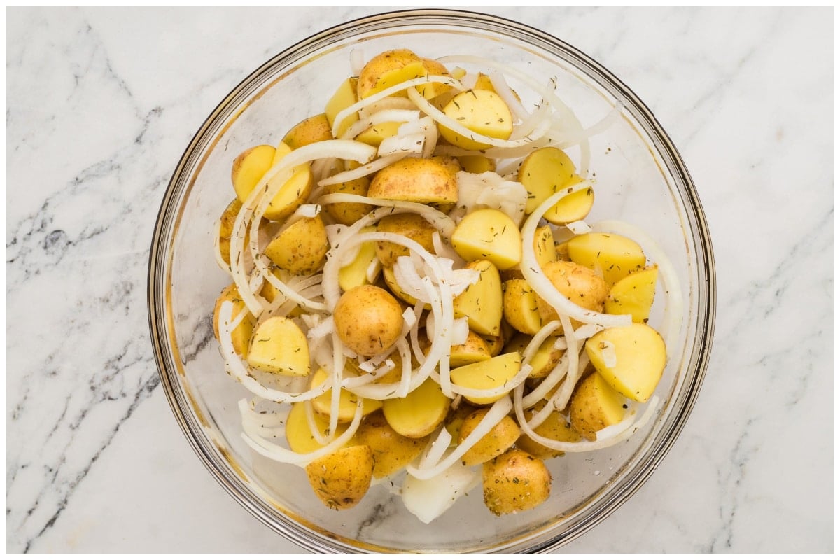 Halved potatoes tossed with onion and some spices and oil on a glass bowl.