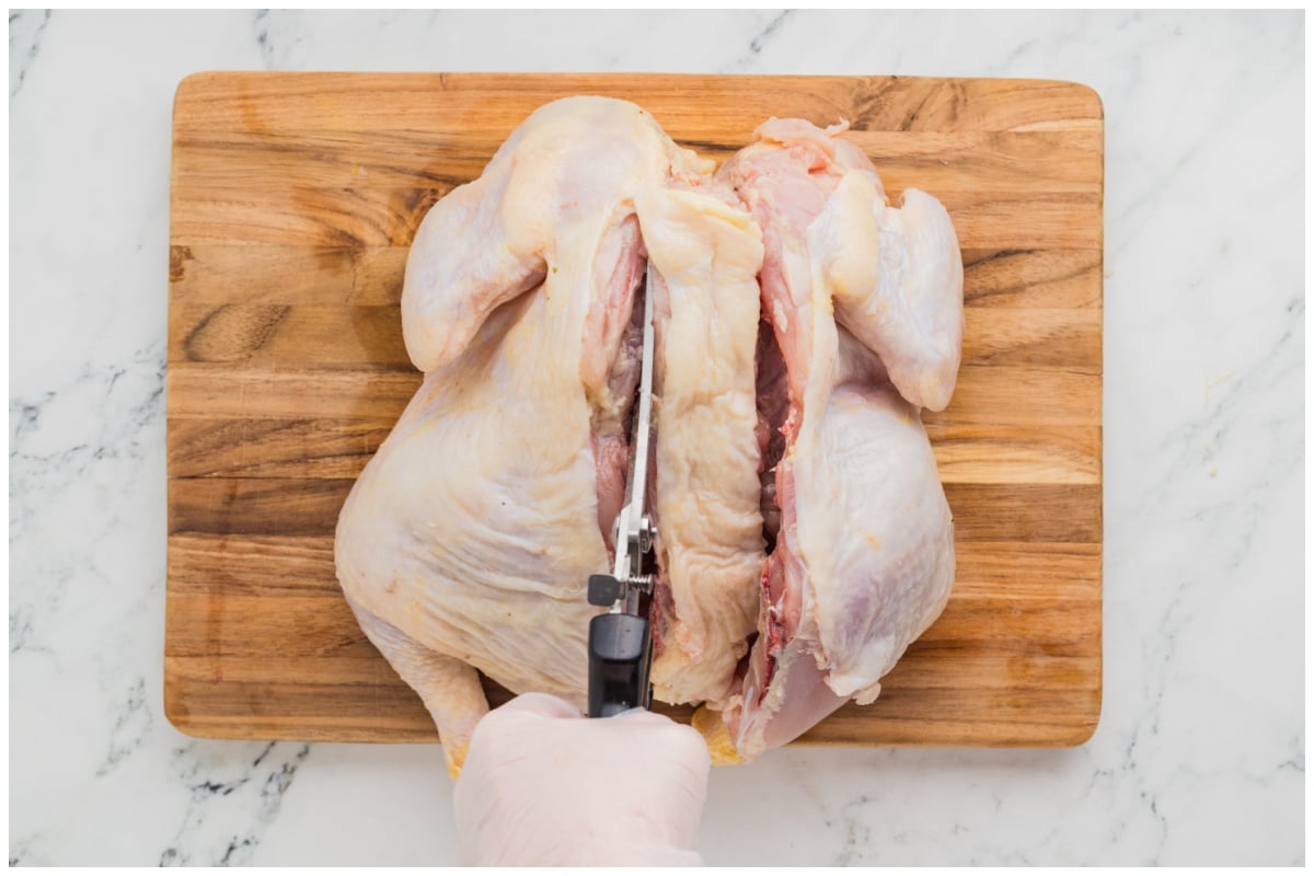 A hand with kitchen sheers cutting the backbone of a whole chicken on a wooden board.