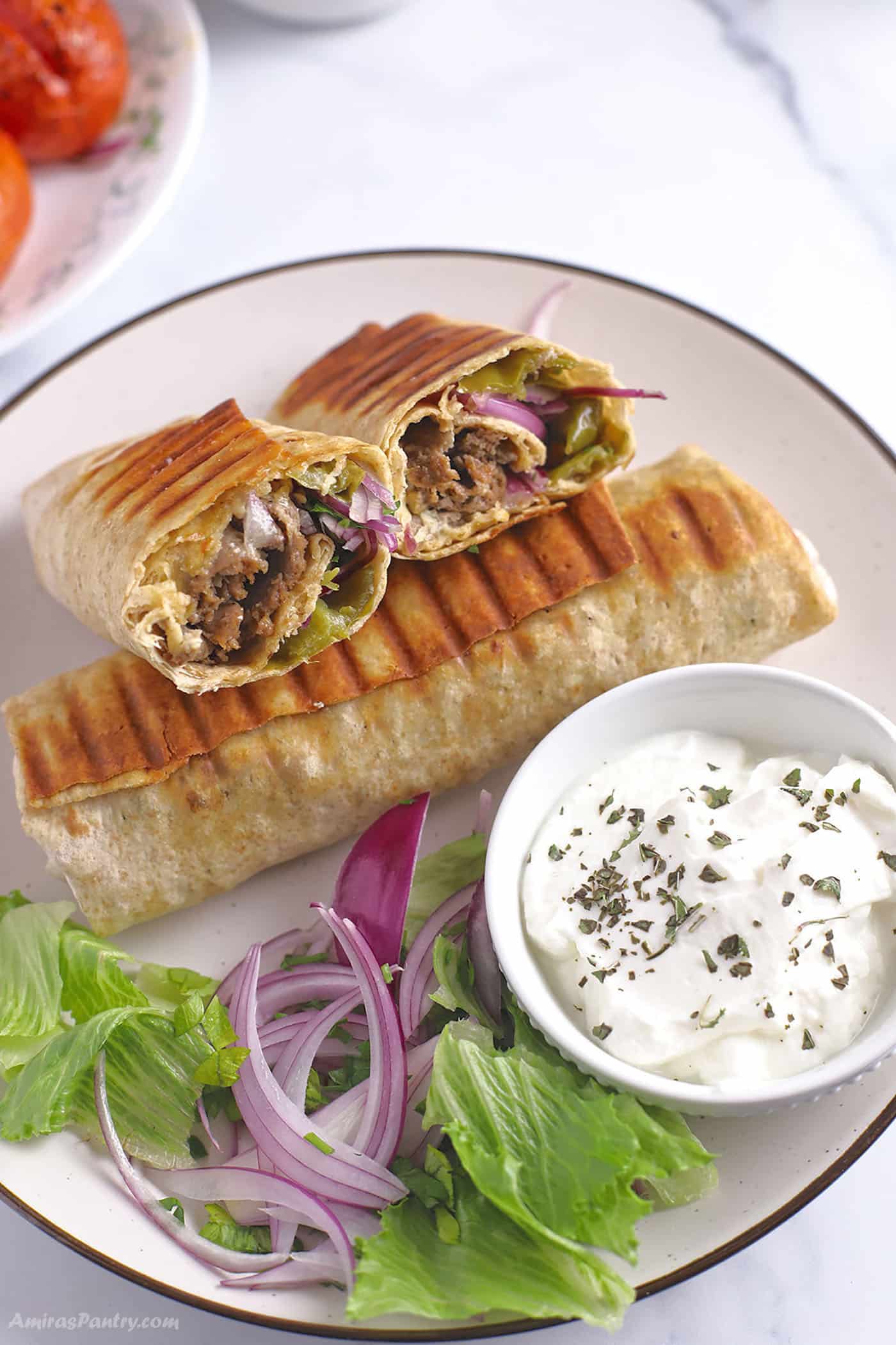 A dinner plae with two Doner Kebab wraps with a bowl of yogurt sauce and greens.