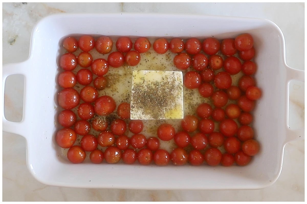 A block of feta cheese in the middle of a baking dish with tomatoes and all drizzeled with oil.