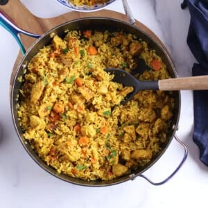 A deep skillet on a wooden board with a spoon lifting some chicken and rice in it.