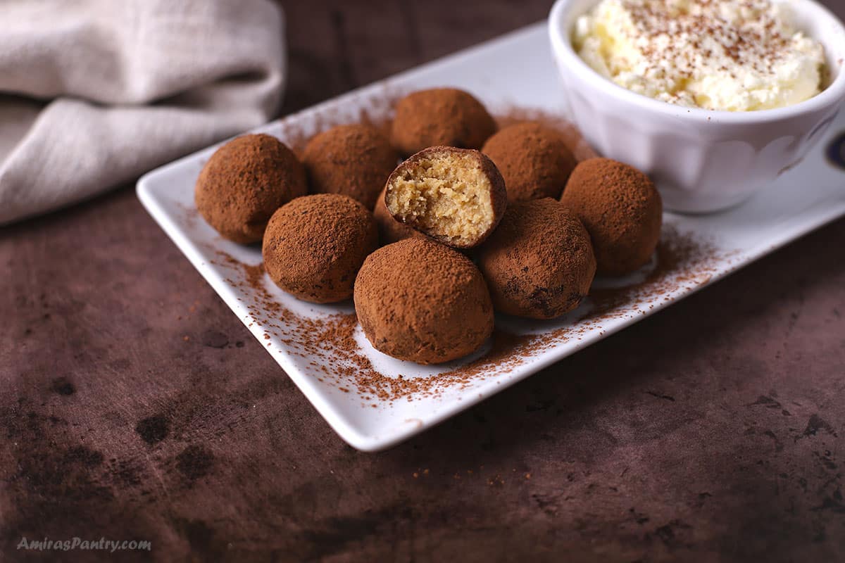 A white serving platter with a pile of tiramisu truffles with a bite taken from one to show texture.