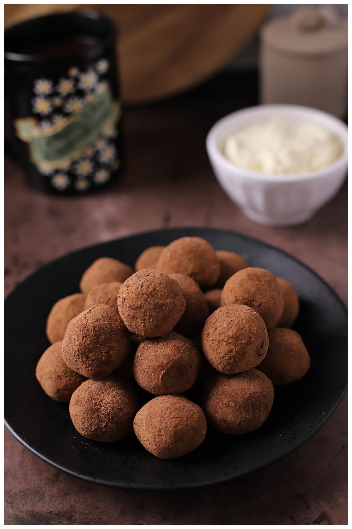A stack of tiramisu balls on a dark plate with coffee cups in the back.