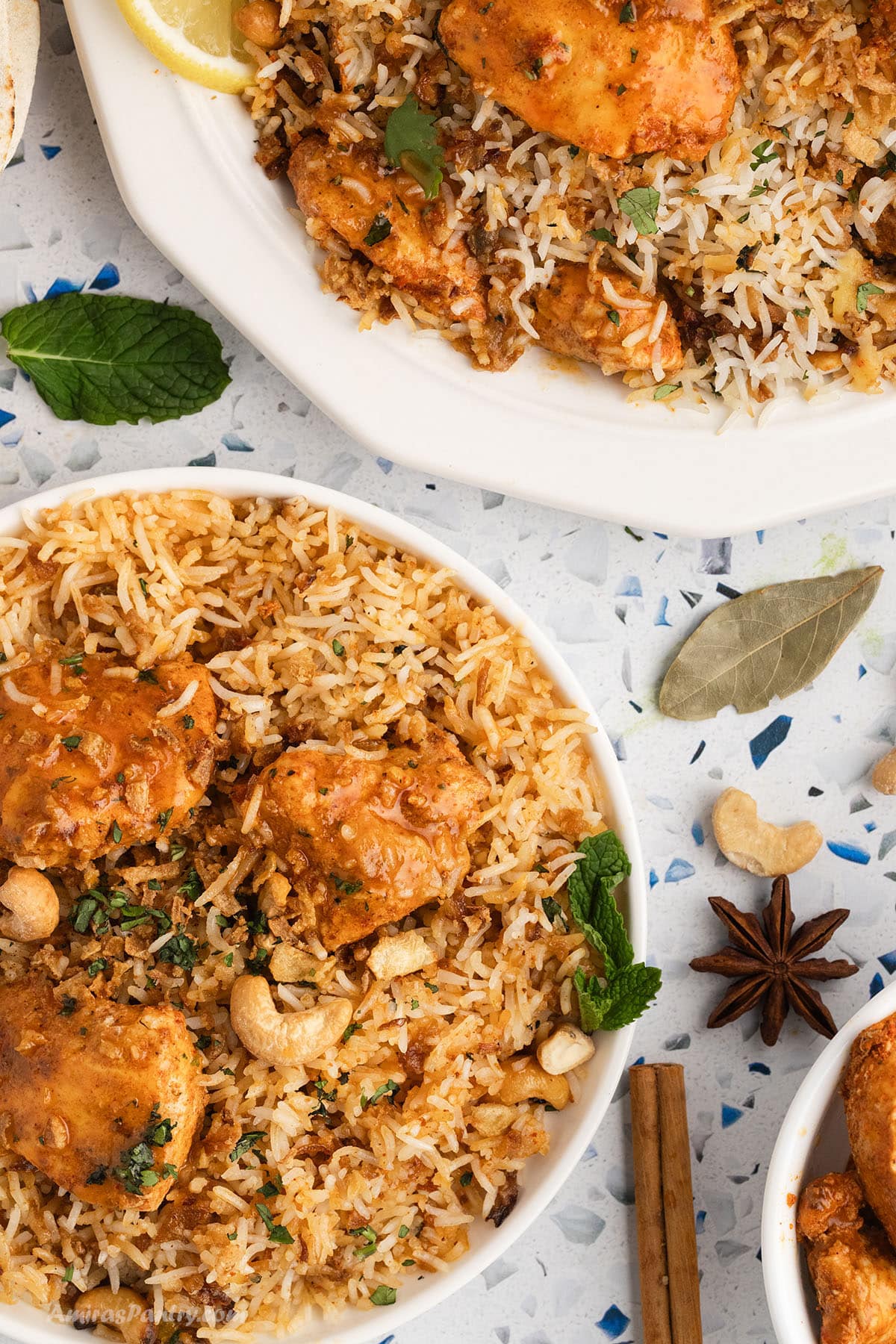 An overhead image of two chicken tikka biryani plates with spices like star anise and cinnamon sticks on the board.
