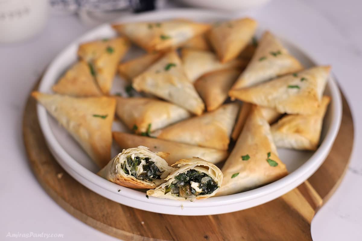 A white platter with spanakopita triangles with one cut in half to show texture.