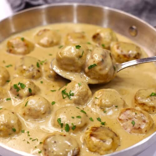 A spoon scooping some chicken Swedish meatballs of a large skillet.