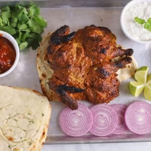 An overhead image of tandoori chicken on a baking sheet with onions, sauces and naan.