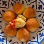Close-up of golden brown Parmesan balls arranged in a blue and white patterned bowl. One ball is open to reveal a soft, cheesy center with a bit of red marinara sauce on the edge.