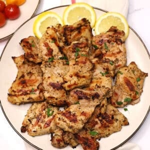 Plate of juicy Italian grilled chicken thighs with charred edges and herbs, garnished with fresh parsley and lemon slices on a light-colored serving platter.