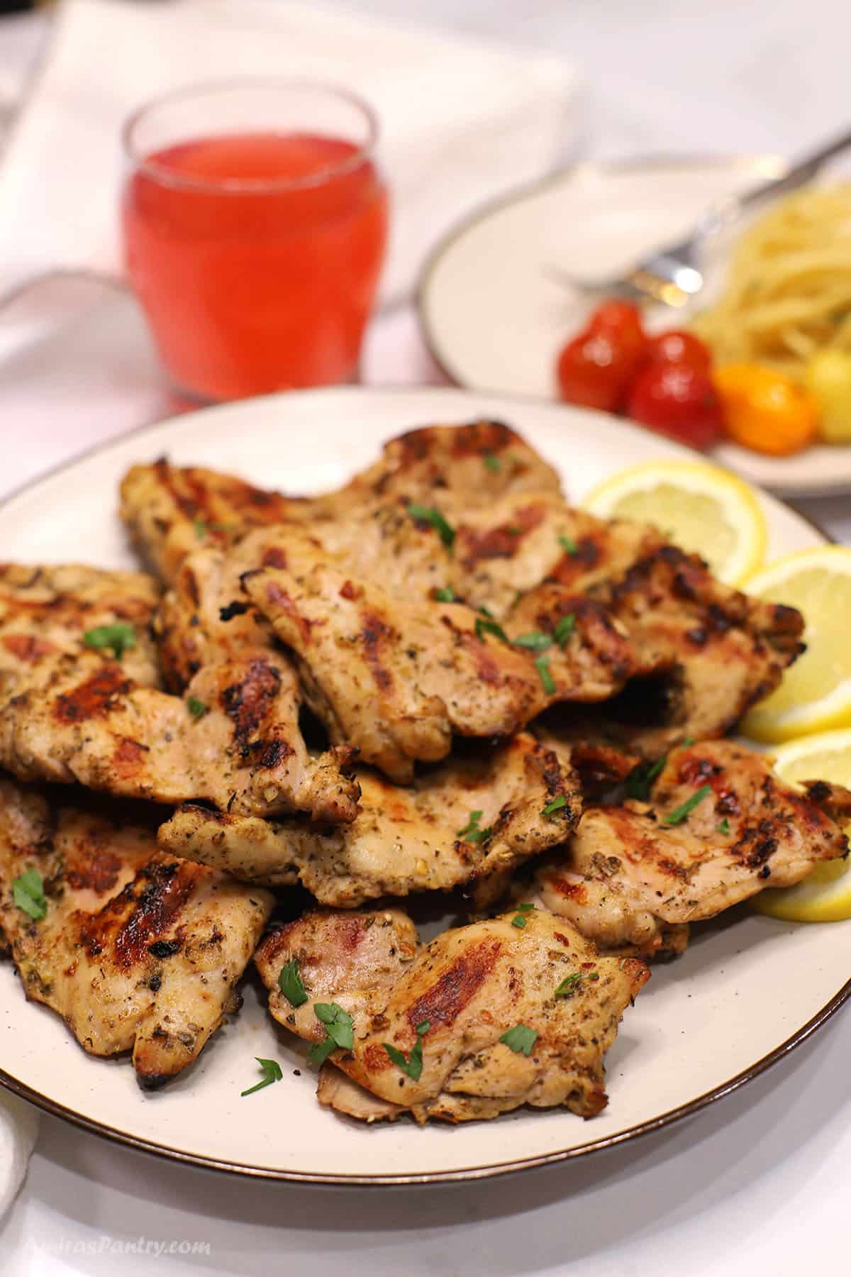 Platter of Italian grilled chicken with crispy charred edges and herbs, garnished with parsley and lemon slices, served with pasta and a pink drink in the background.