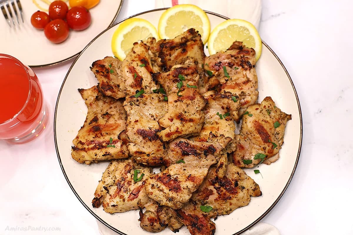 Overhead view of grilled Italian grilled chicken with herbs, served on a platter with lemon slices, cherry tomatoes, and a pink drink on the side.
