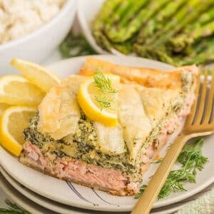 Angled close-up of Salmon Wellington served on stacked plates, showing layers of flaky puff pastry, juicy baked salmon, and rich creamy spinach filling.