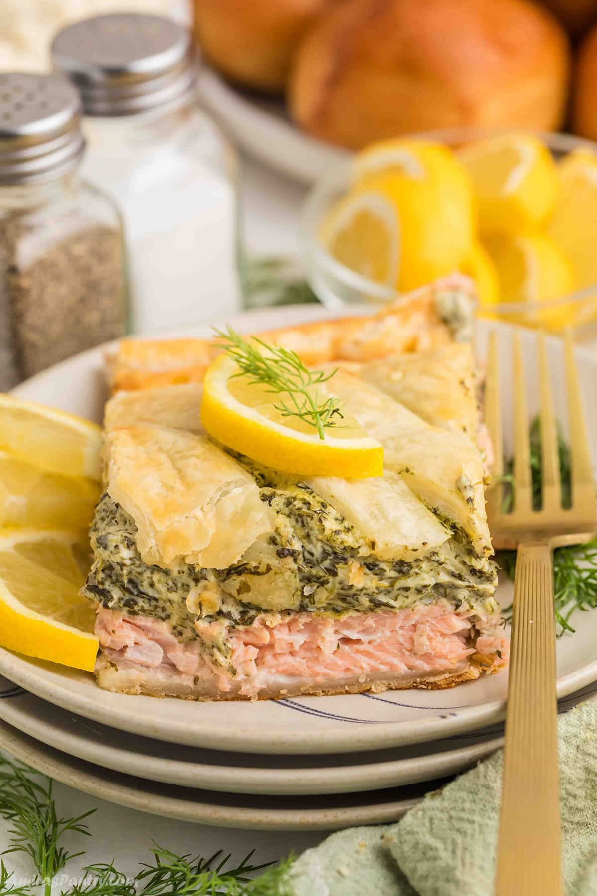 Close up of a slice of Salmon Wellington on stacked plates, featuring flaky golden puff pastry layered with tender pink salmon and a creamy spinach filling, garnished with lemon slices and fresh dill, with a gold fork and dinner rolls blurred in the background.
