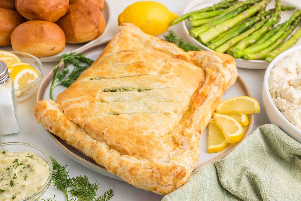 Whole baked Salmon Wellington with golden flaky puff pastry served on a platter with lemon slices, roasted asparagus, mashed potatoes, fresh dill, and dinner rolls, styled as an elegant meal presentation.