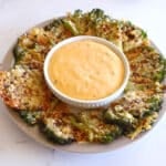 An overhead shot of a white plate filled with crispy smashed broccoli florets arranged in a circle around a small white bowl of creamy orange dipping sauce.
