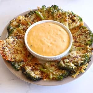 An overhead shot of a white plate filled with crispy smashed broccoli florets arranged in a circle around a small white bowl of creamy orange dipping sauce.