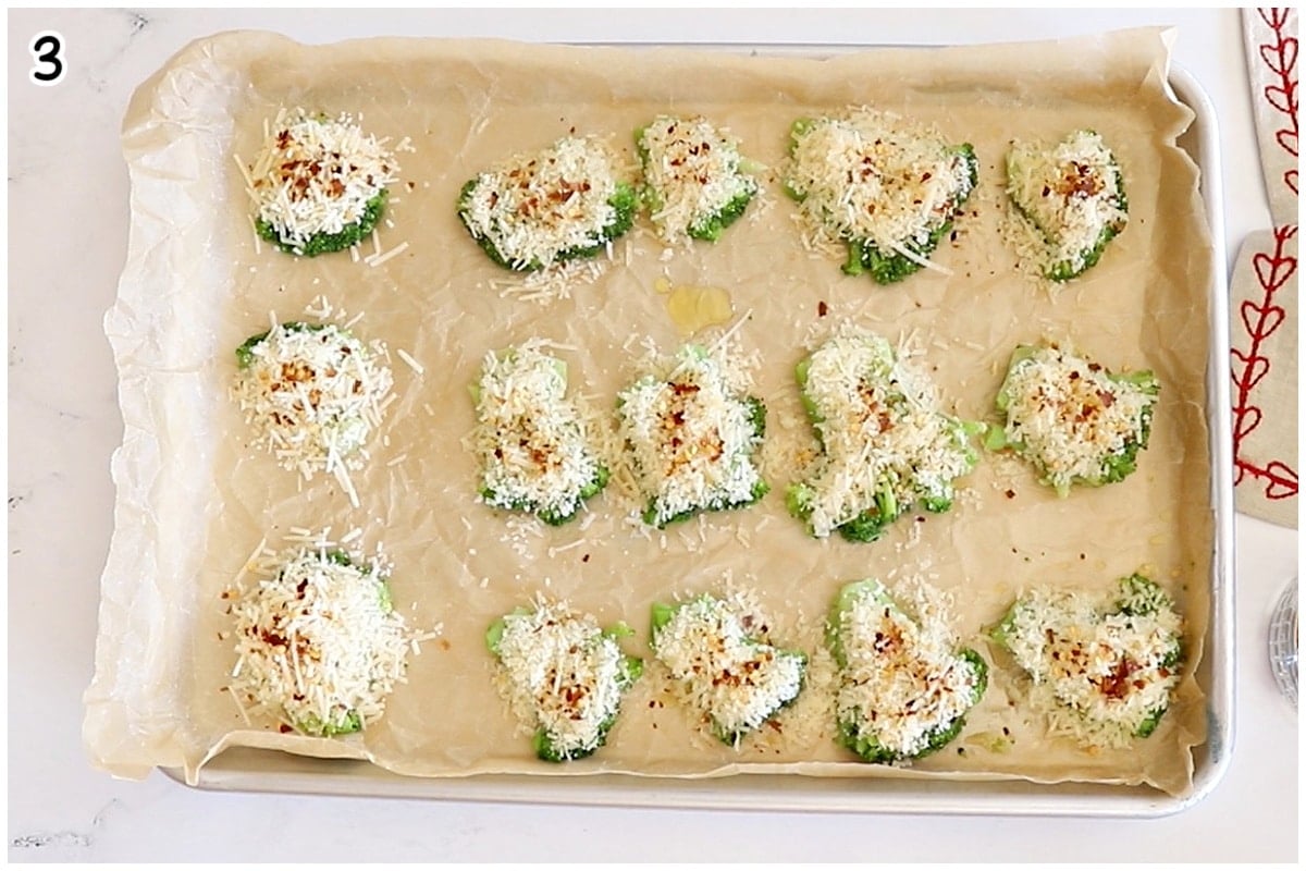 An overhead view of a large baking sheet with rows of flattened broccoli florets covered with shredded cheese and red pepper flakes.