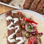 Close-up of two grilled kofta kebabs drizzled with creamy tahini sauce are served on a soft flatbread with a fresh tomato salad. A tray of additional kebabs in the background adds context to this colorful and hearty Middle Eastern-inspired meal.