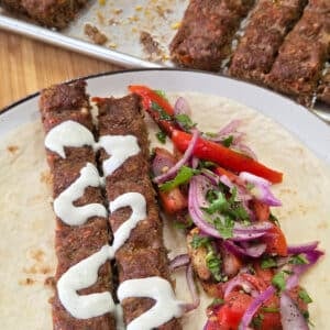 Close-up of two grilled kofta kebabs drizzled with creamy tahini sauce are served on a soft flatbread with a fresh tomato salad. A tray of additional kebabs in the background adds context to this colorful and hearty Middle Eastern-inspired meal.