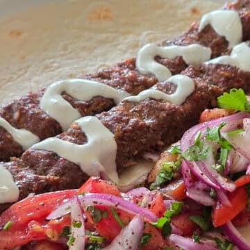 Close-up of grilled kofta kebabs drizzled with creamy tahini sauce, served alongside a fresh tomato, red onion, and parsley salad.