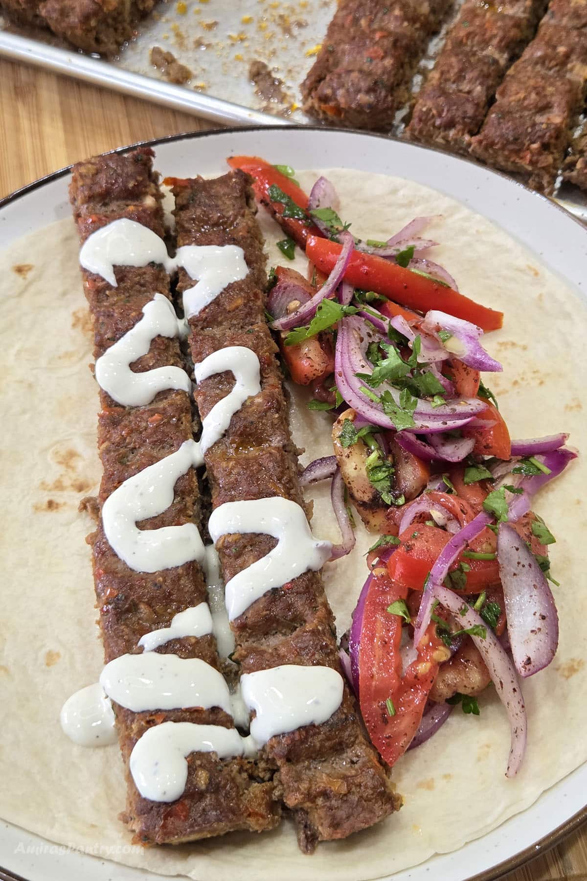 Two grilled Adana kebabs drizzled with creamy garlic sauce are served on a soft flatbread with a fresh tomato, red onion, and parsley salad.