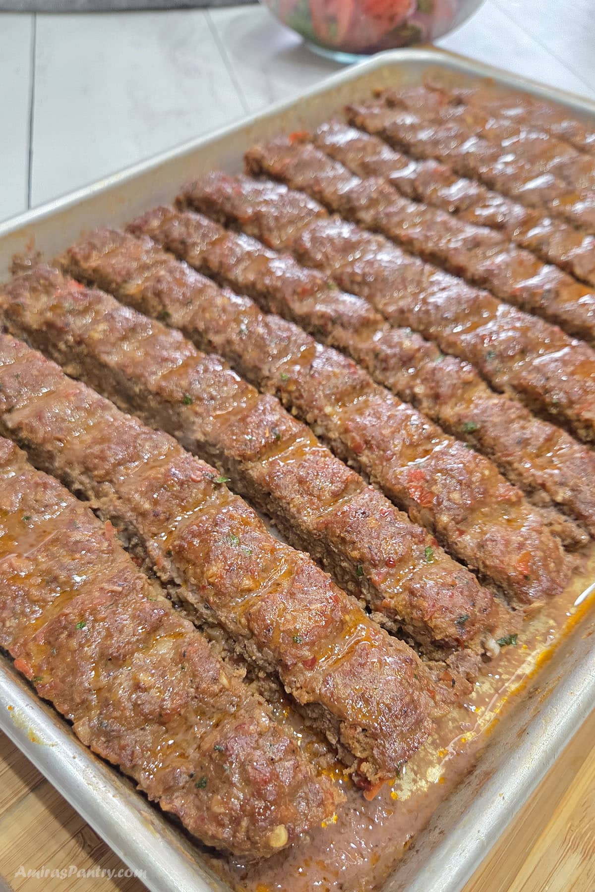 Sheet pan of freshly baked Adana kebabs arranged in neat rows, glistening with flavorful juices and rendered fat. The golden-brown ground meat kebabs are scored into portions and ready to serve, with a bowl of fresh salad blurred in the background.