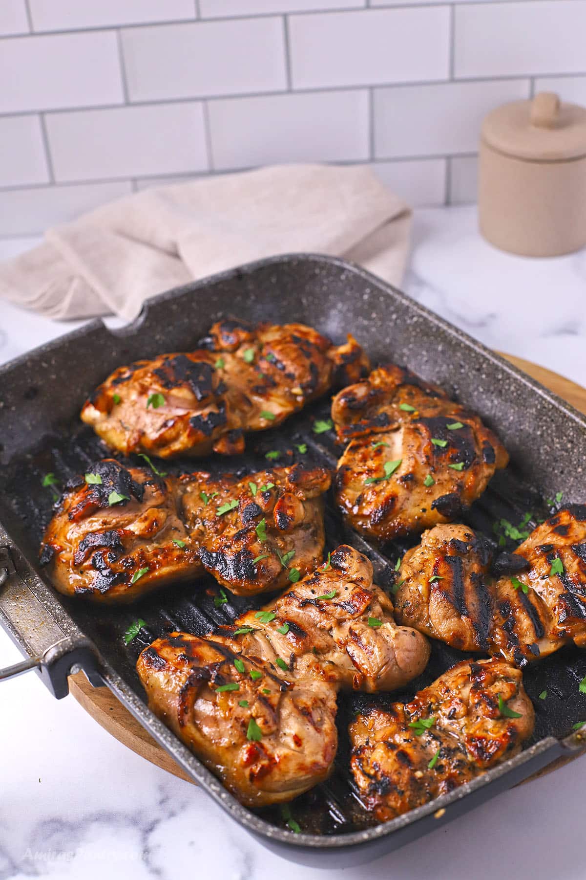 Grilled balsamic chicken thighs with charred grill marks and a sprinkle of fresh parsley, served in a dark square grill pan on a marble countertop.