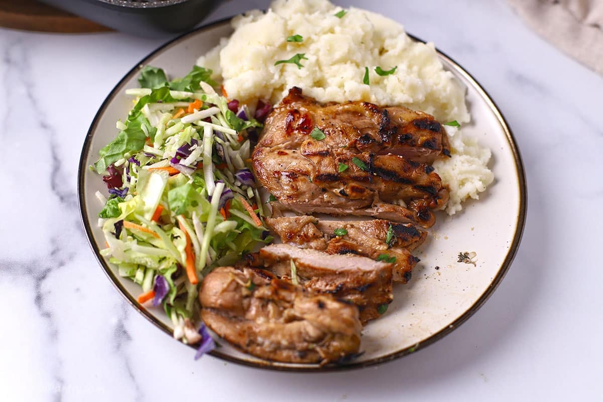 A white plate featuring sliced, grilled balsamic chicken thighs with deep char marks, served alongside a mound of fluffy mashed potatoes sprinkled with parsley and a crisp green slaw with shredded carrots and purple cabbage.