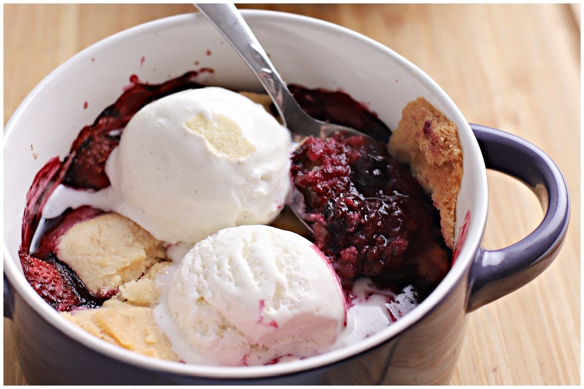 Close-up angled shot of baked berry cobbler with golden biscuit topping in a blue handled dish, topped with two melting scoops of vanilla ice cream and a spoon.