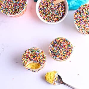 Overhead view of Dot Cakes in glass jars topped with rainbow sprinkles, one cut into showing yellow cake and frosting layers, with a spoon holding a bite.