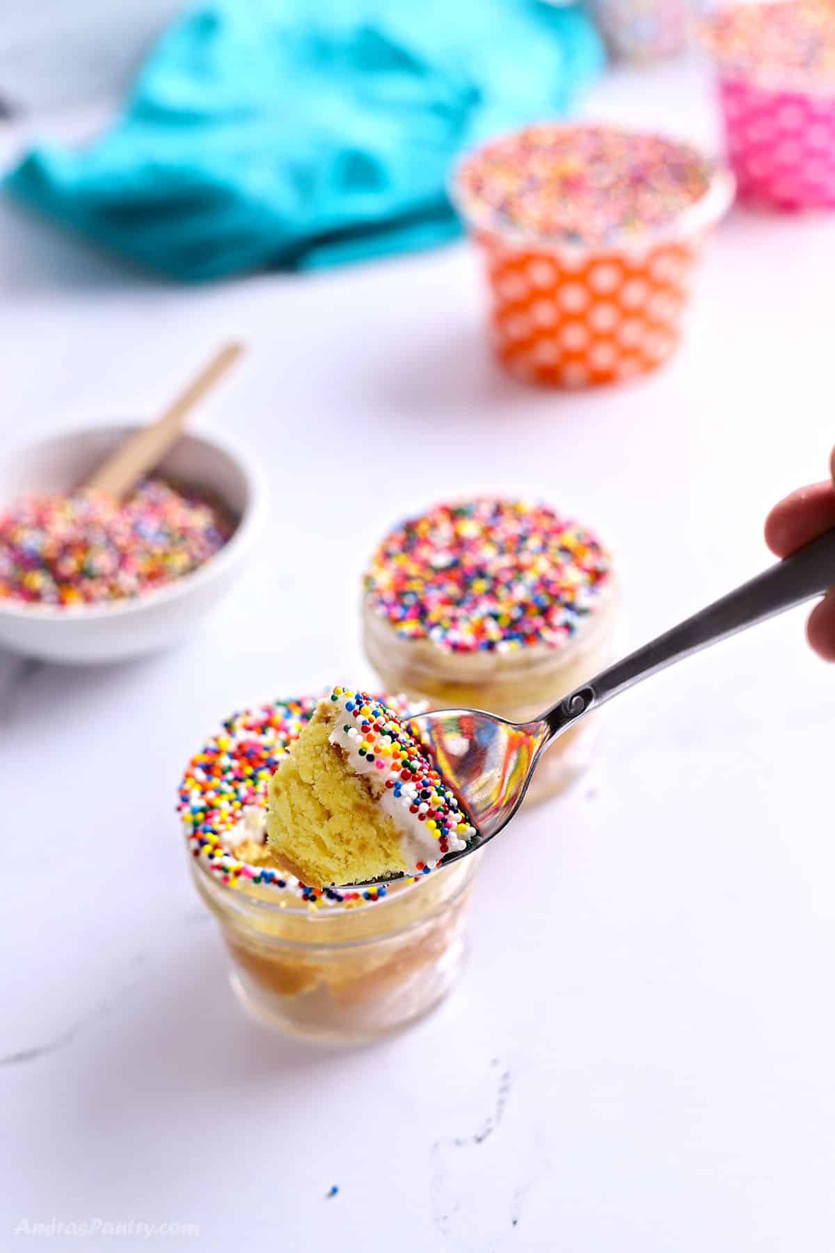 A spoon scooping a bite of yellow cake layered with frosting from a Dot Cake in a glass jar, topped with rainbow sprinkles.