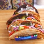 A row of four falafel tacos in toasted pita bread, held upright in a colorful striped taco rack on a wooden cutting board.