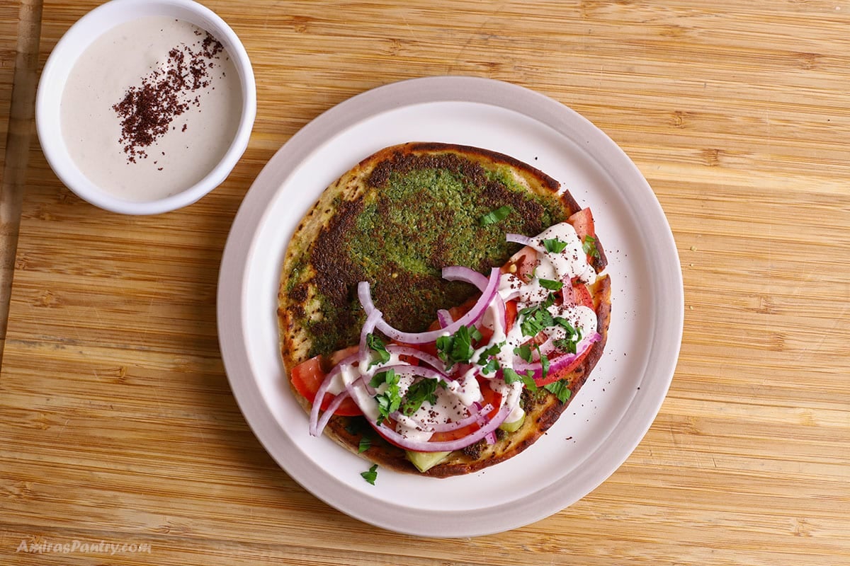 Open-faced falafel taco on a white plate, topped with tomatoes, red onion, parsley, tahini drizzle, and sumac, with a small bowl of tahini on the side.